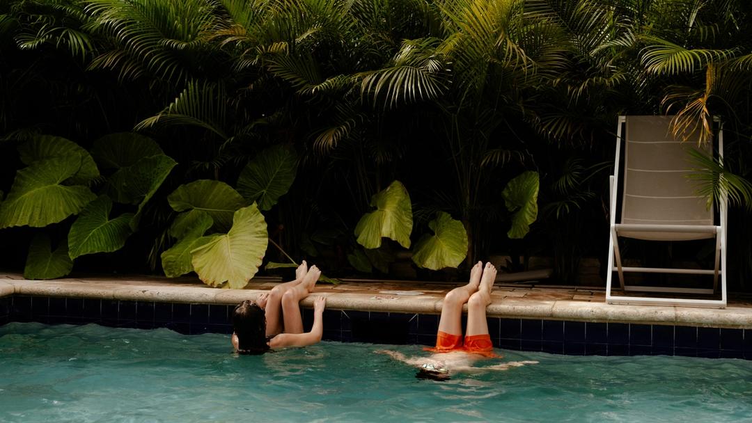 Two people swimming in a pool with lush greenery behind them.