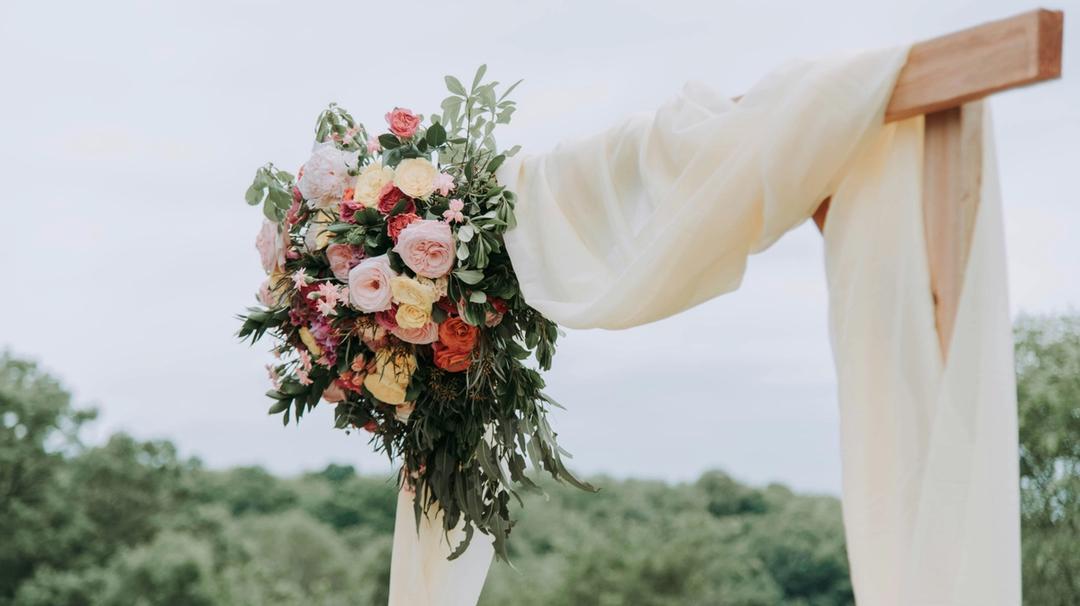 Floral wedding arrangement attached to a wooden ceremony arch outdoors. 