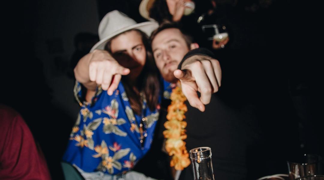 A group of men sitting at a table with drinks pointing at the camera.