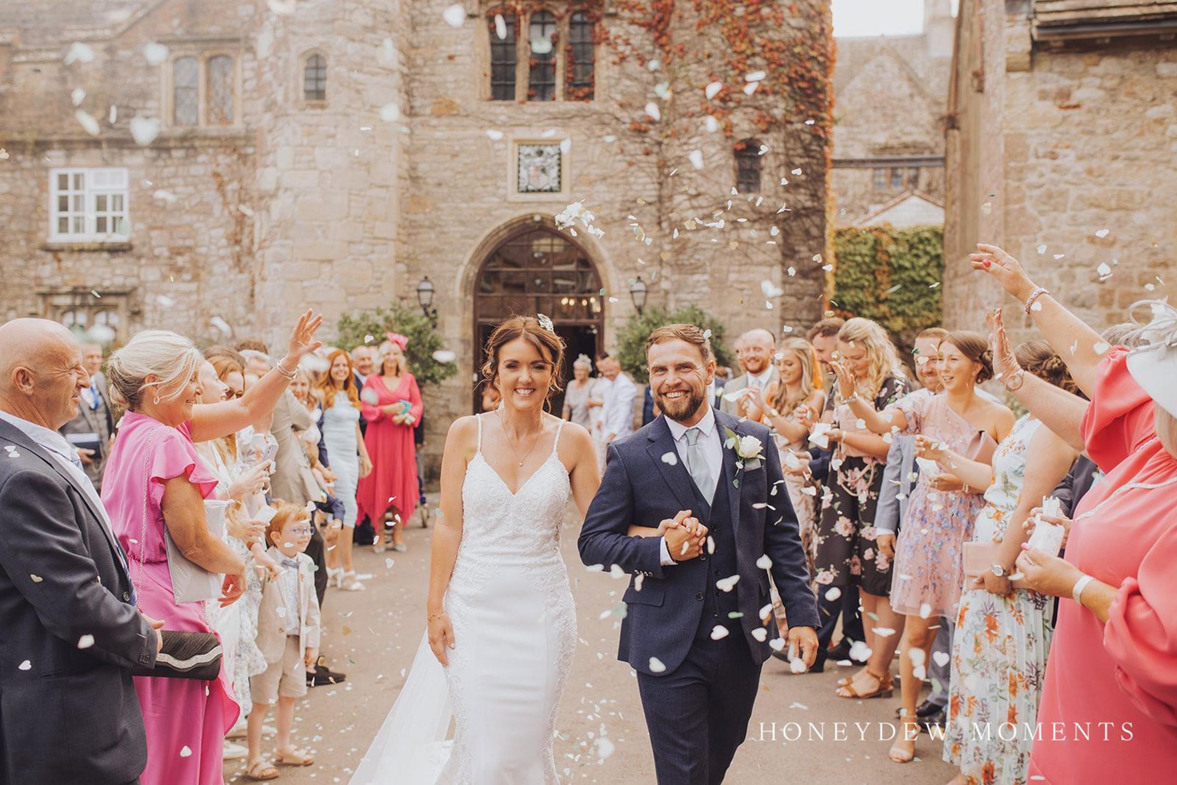 Newlyweds walking through a confetti toss outside a historic stone gatehouse