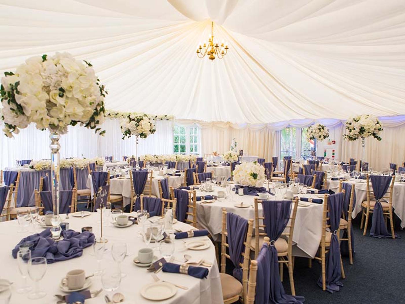 Marquee reception laid with round tables, blue chair sashes and tall floral centrepieces