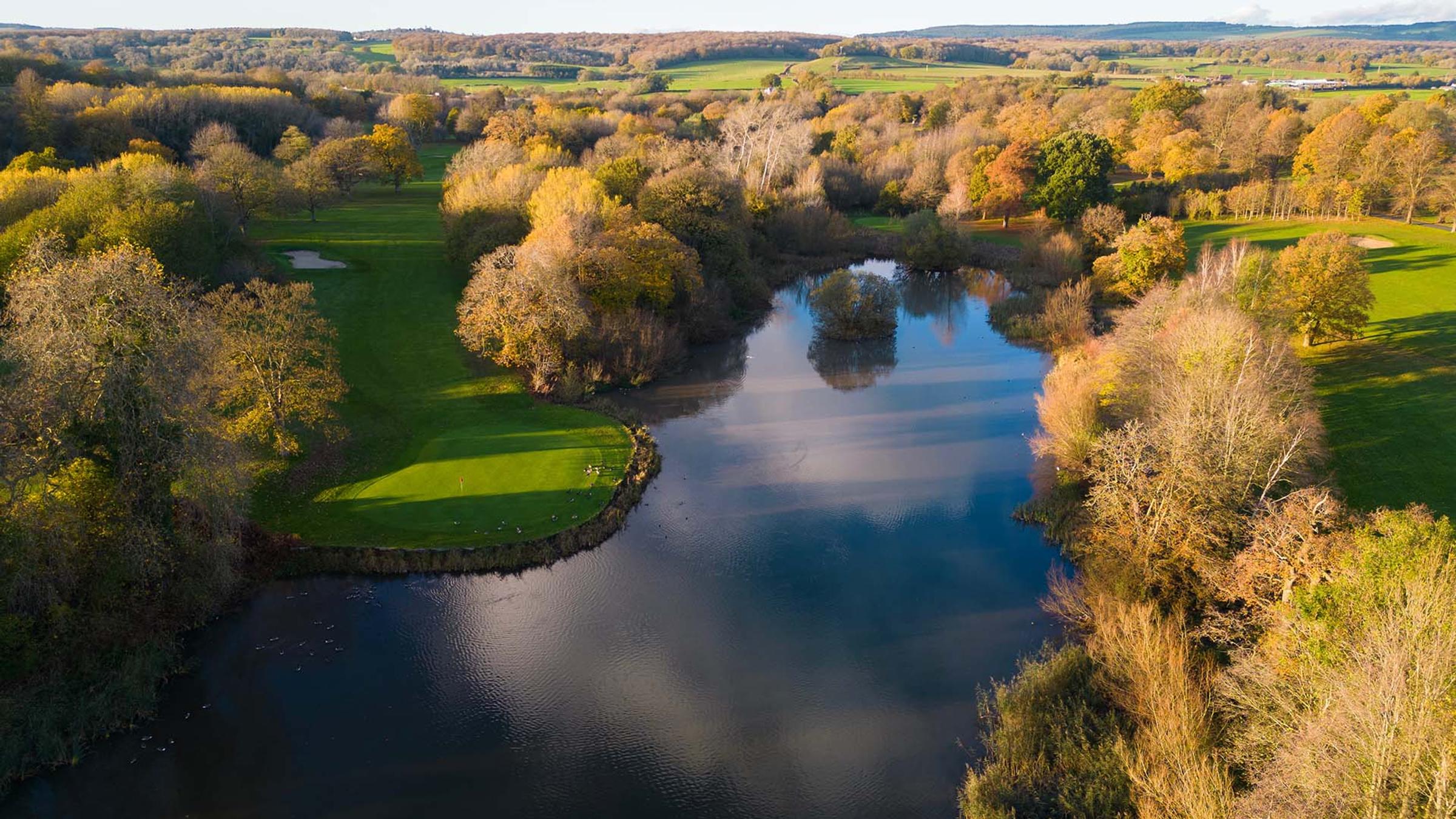 Aerial view of a golf green beside a lake, surrounded by autumn woodland