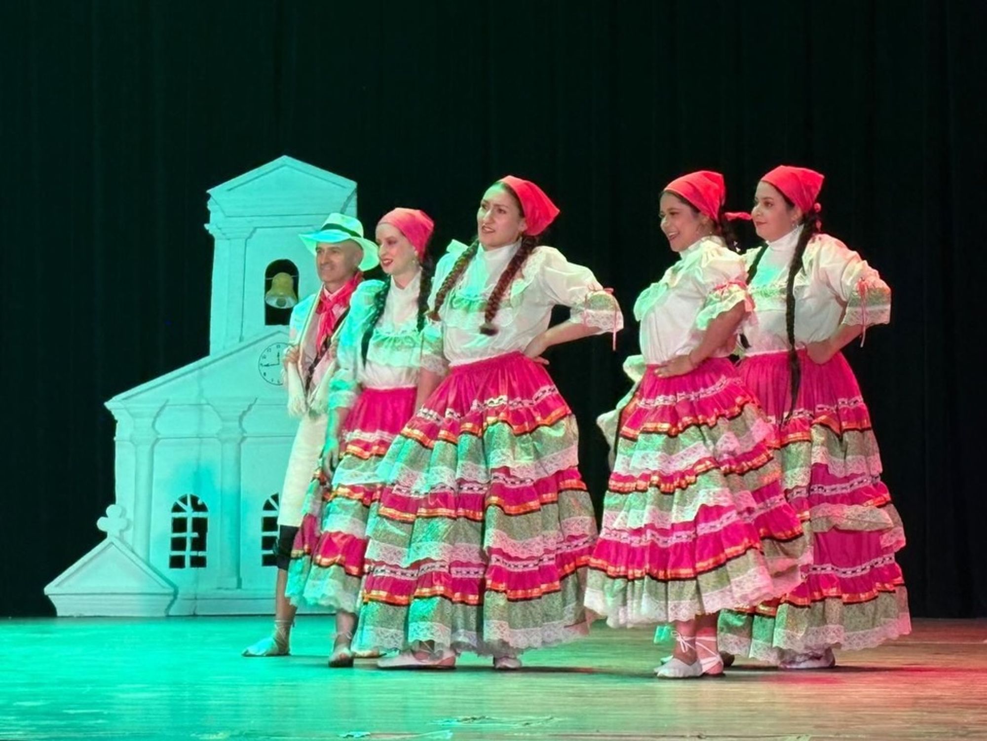 Bailarinas con traje campesino y pañoleta rosada frente a una capilla de utilería
