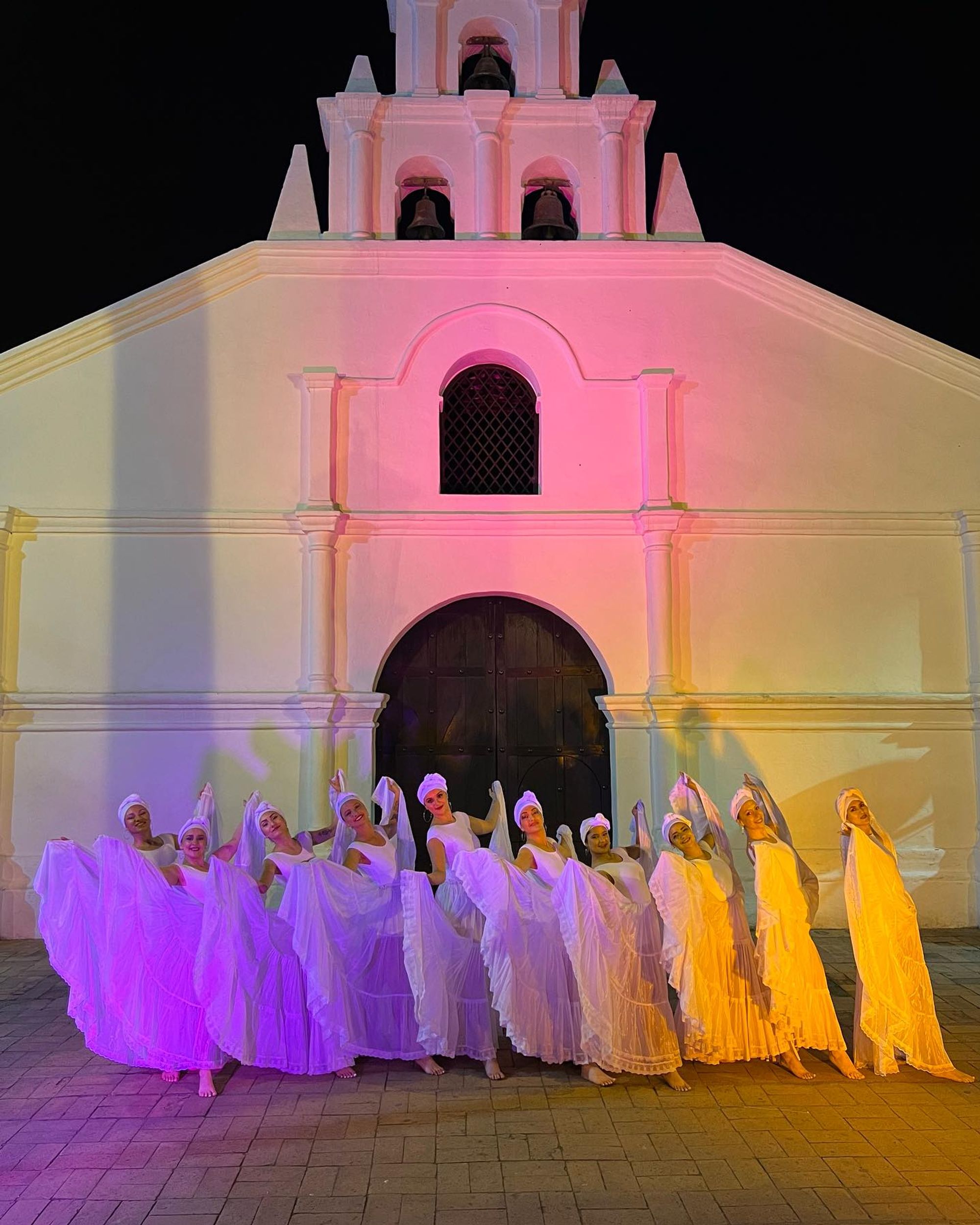 Bailarinas vestidas de blanco frente a una capilla iluminada de noche