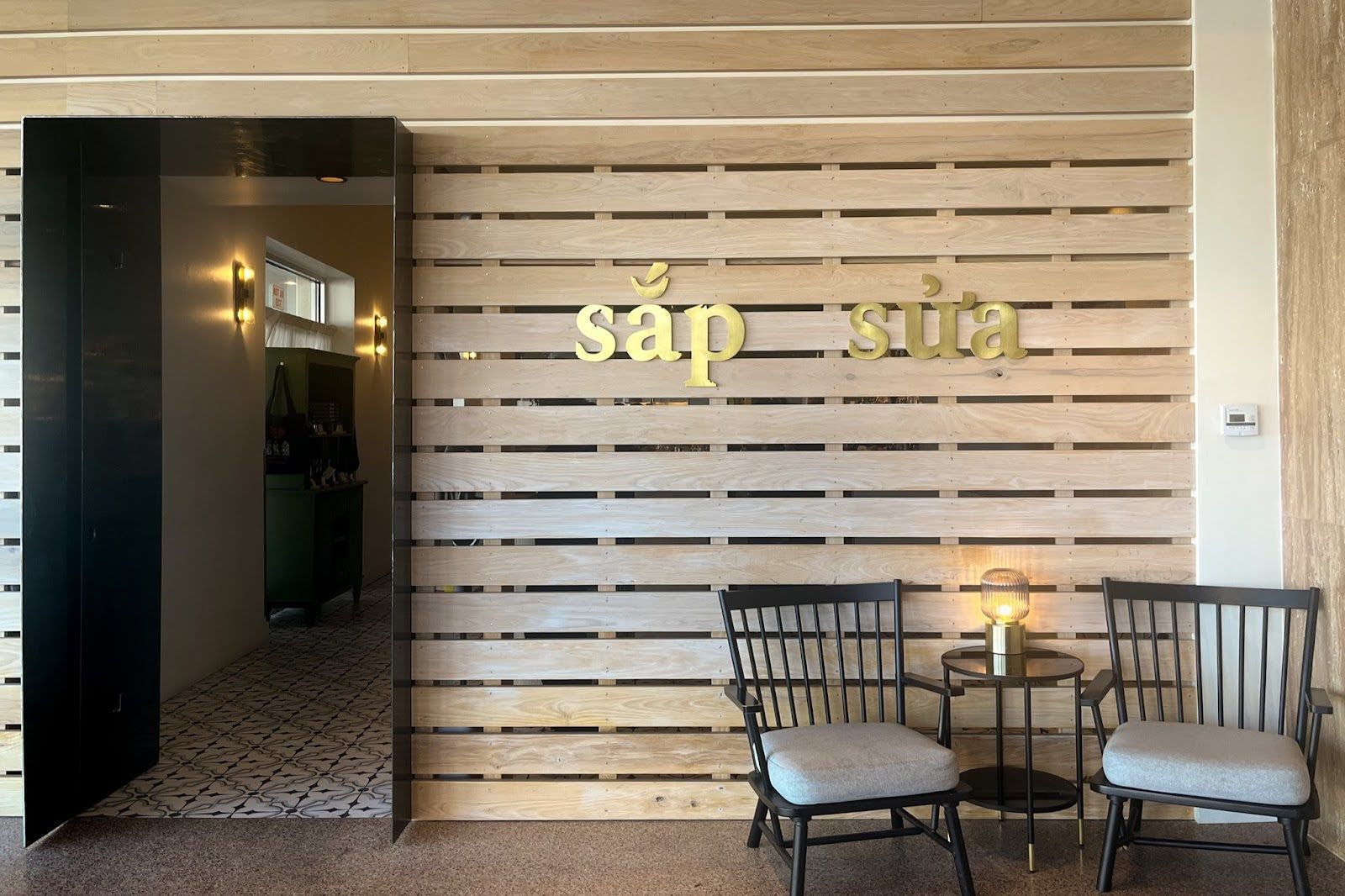 A modern entrance with a wooden slat wall featuring "sắp sữa" in gold letters, two chairs, a table with a lamp, and a dark doorway.