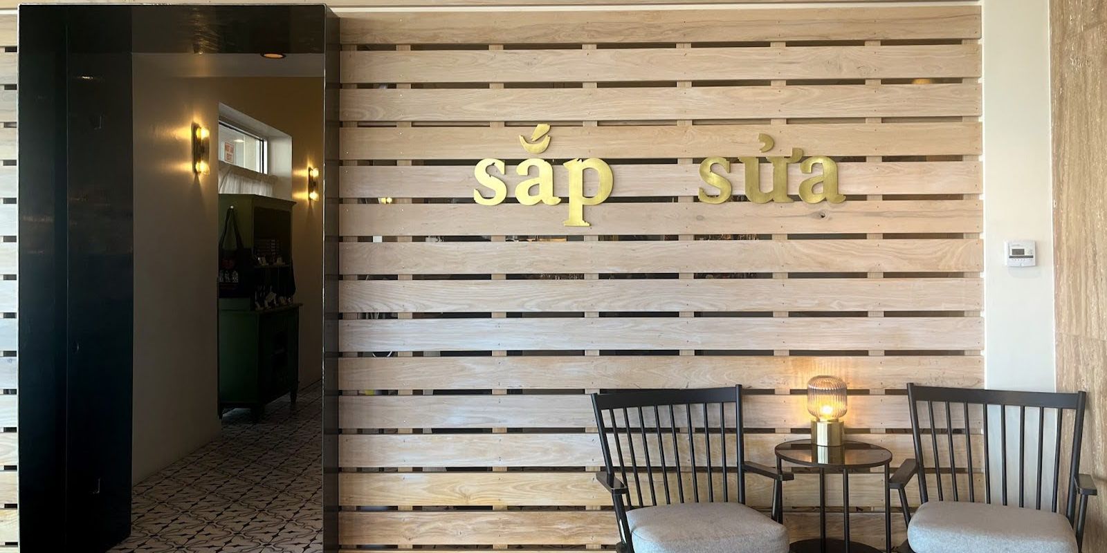 A modern entrance with a wooden slat wall featuring "sắp sữa" in gold letters, two chairs, a table with a lamp, and a dark doorway.