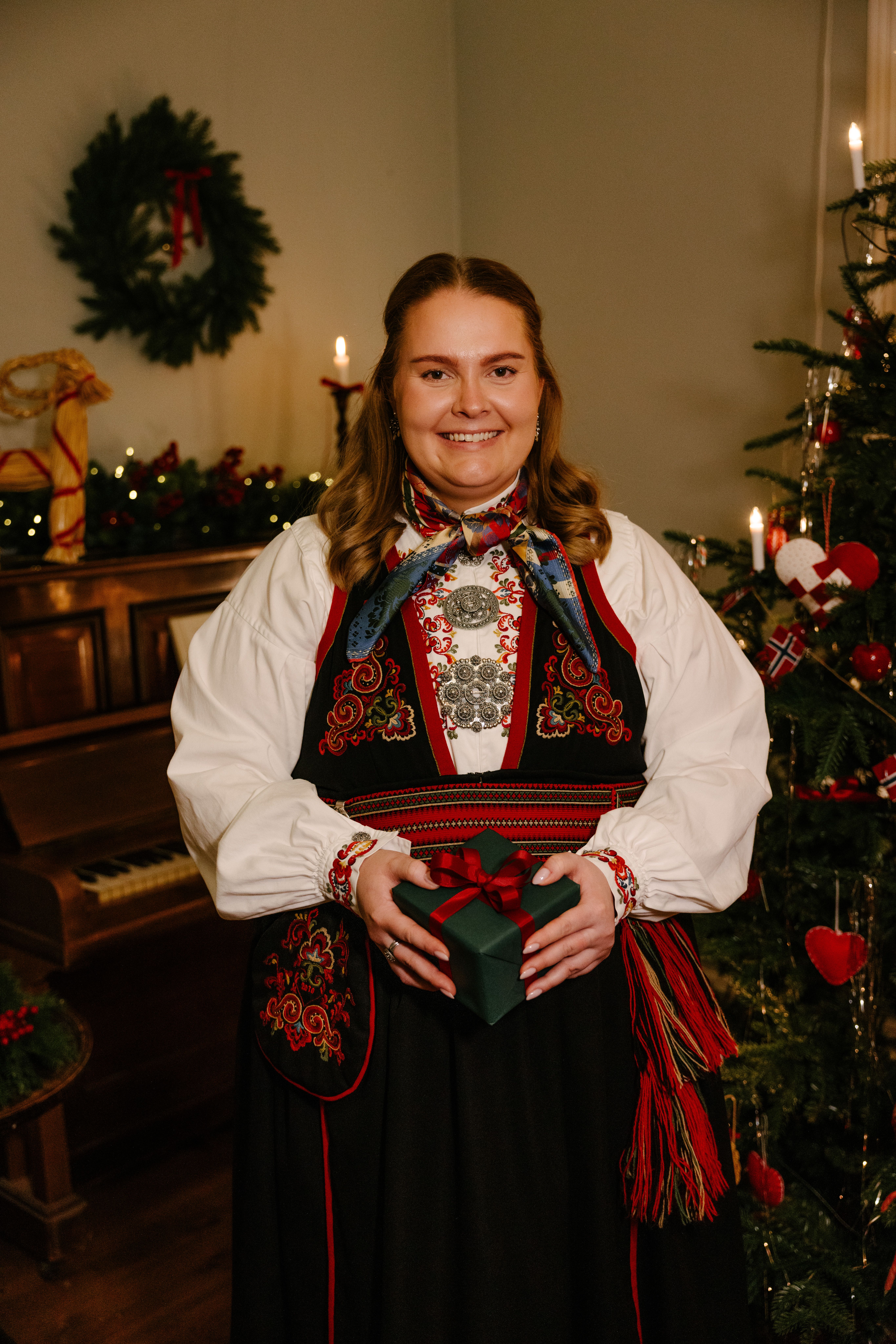 Kvinne i bunad som holder en grønn gave foran et juletre