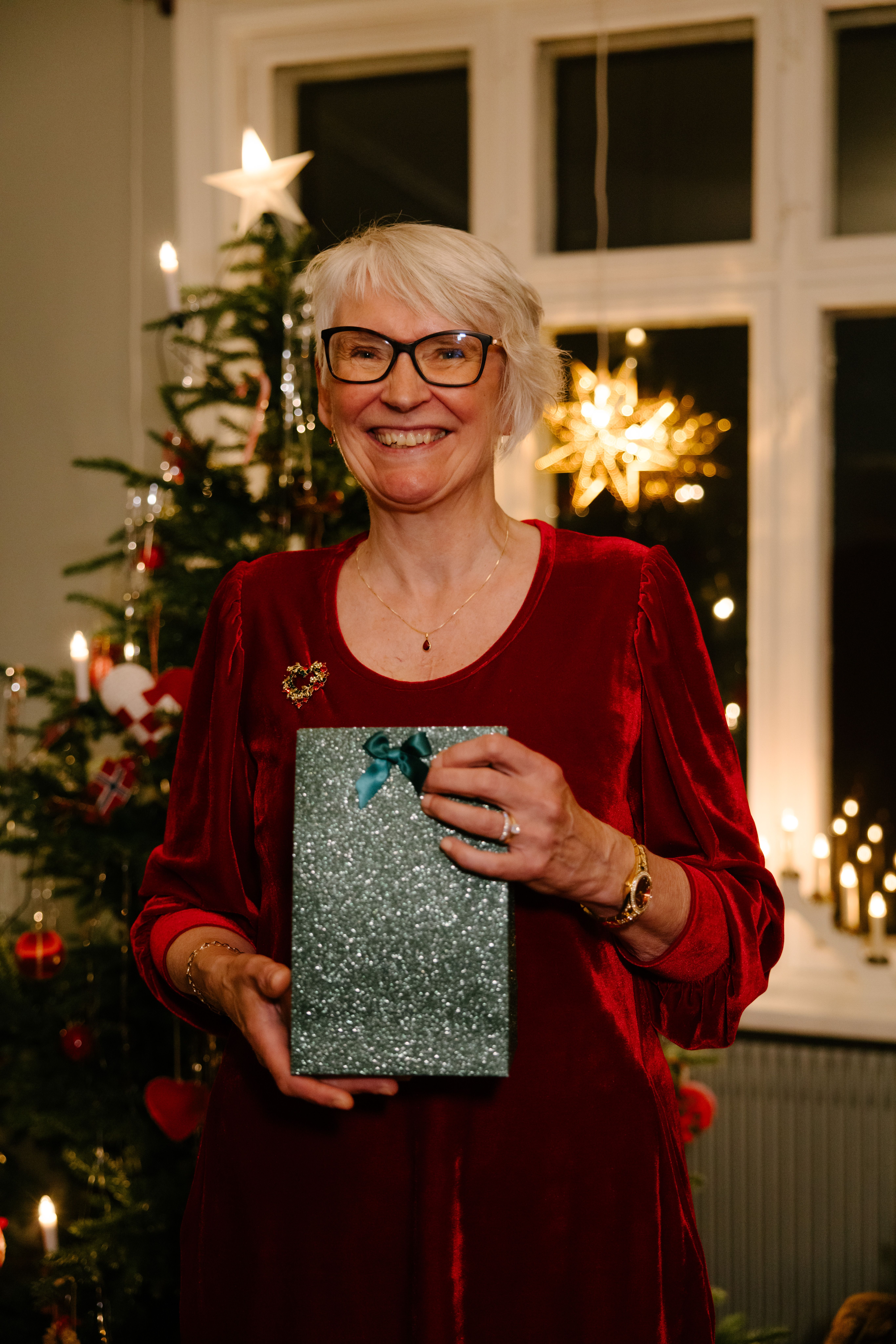 Kvinne som smiler i rød kjole med en grønn gavepakke og juletre i bakgrunn