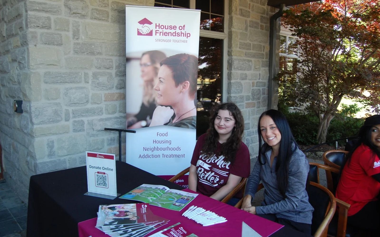 Two House of Friendship team members sit at an information table at the Jackman Charity Golf Tournament, in front of a Stronger Together banner.