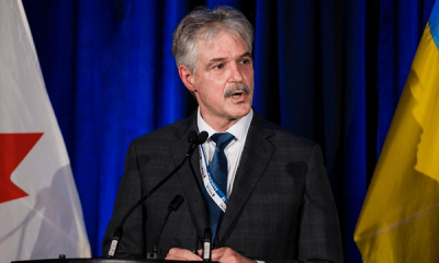 Stephen Matier, the founder and CEO of Maritime Launch Services, at a press conference with flags behind him against a blue curtain wearing a suit and navy tie