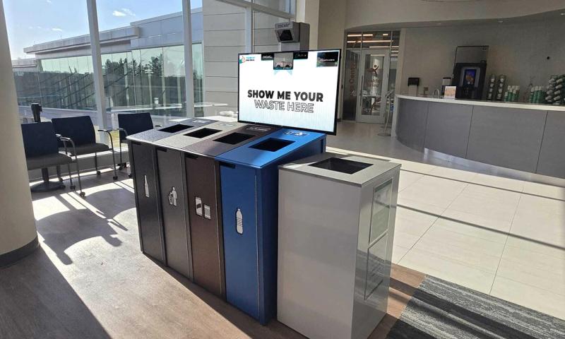 Oscar's screen reading "Show me your waste here" sits under a camera and above a set of trash and recycling bins at an airport