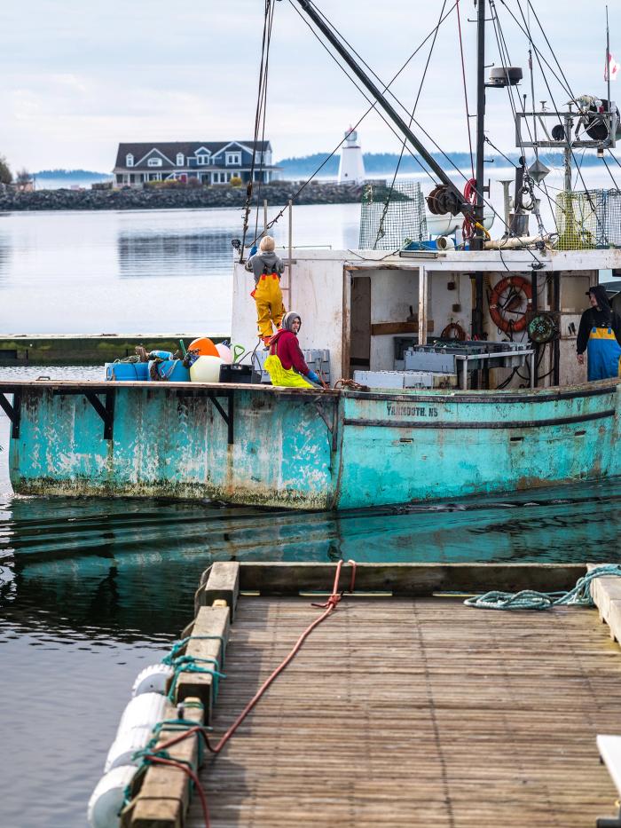 A turquoise blue lobster and crab fishing vessel with the name All Done and a crew of fishers working on it pulls out of the wharf filled with ropeless fishing gear near St. Andrews New Brunswick