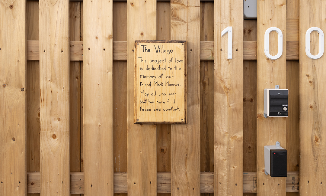 A wood plaque on a wood wall that reads "The village this project of love is dedicated to the memory of our friend Mark Munroe May all who seek shelter here find peace and comfort" next to address numbers that read "100" and two black panels