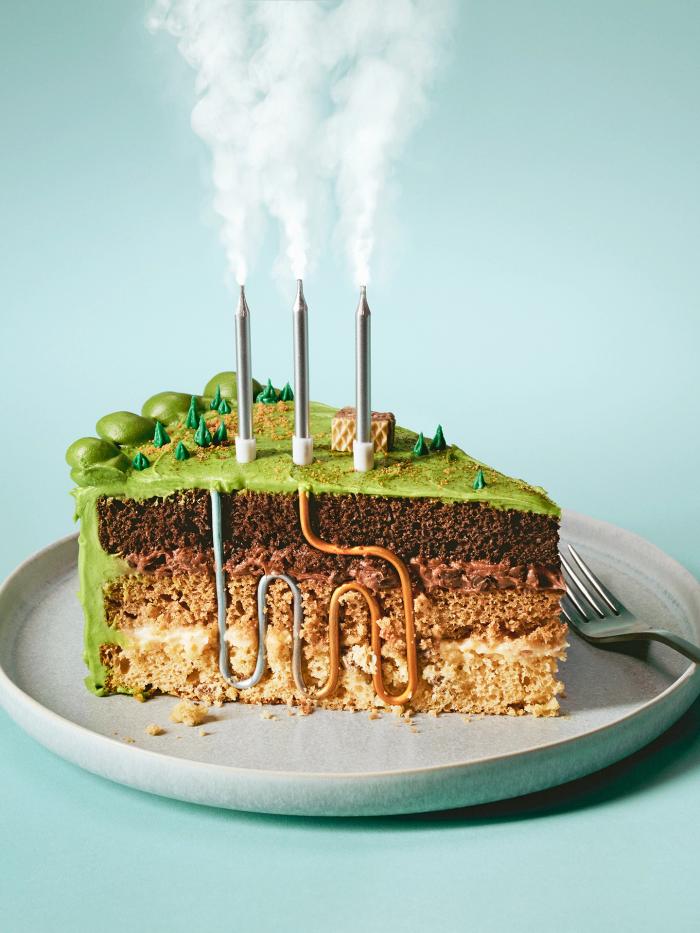 A slice of cake with green icing designed to look like layers of the Earth’s crust, with silver candles representing steam and a series of pipes winding through the layers of the cake to illustrate a piece on next-gen geothermal energy in Canada.