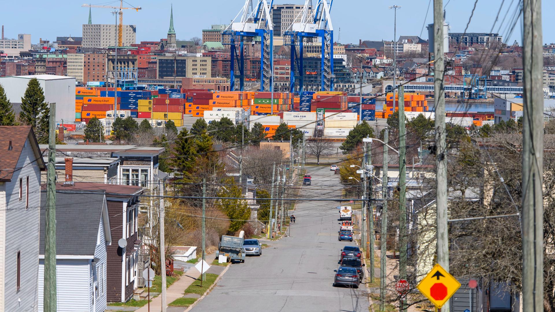 A photo illustration of a cargo ship with the Hapag-LLoyd name on the site and a crane behind it with people walking in the foreground across the water at the Port of Saint John