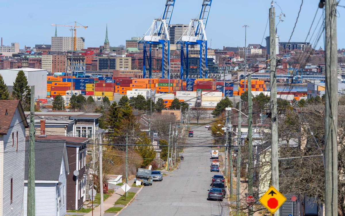 A photo illustration of a cargo ship with the Hapag-LLoyd name on the site and a crane behind it with people walking in the foreground across the water at the Port of Saint John