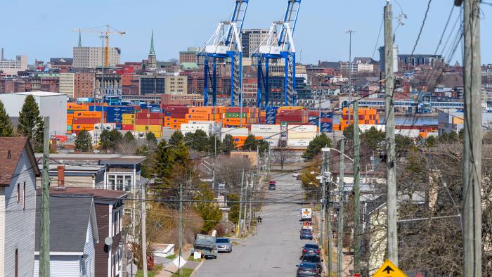 A photo illustration of a cargo ship with the Hapag-LLoyd name on the site and a crane behind it with people walking in the foreground across the water at the Port of Saint John