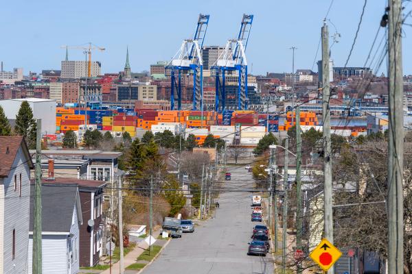 A photo illustration of a cargo ship with the Hapag-LLoyd name on the site and a crane behind it with people walking in the foreground across the water at the Port of Saint John