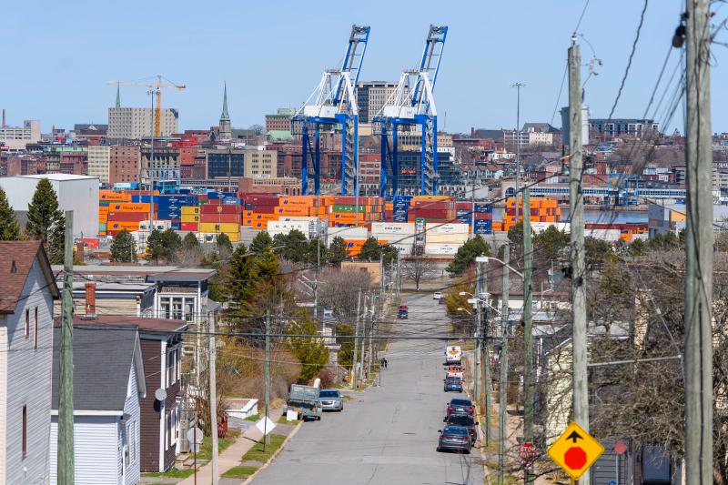 A photo illustration of a cargo ship with the Hapag-LLoyd name on the site and a crane behind it with people walking in the foreground across the water at the Port of Saint John