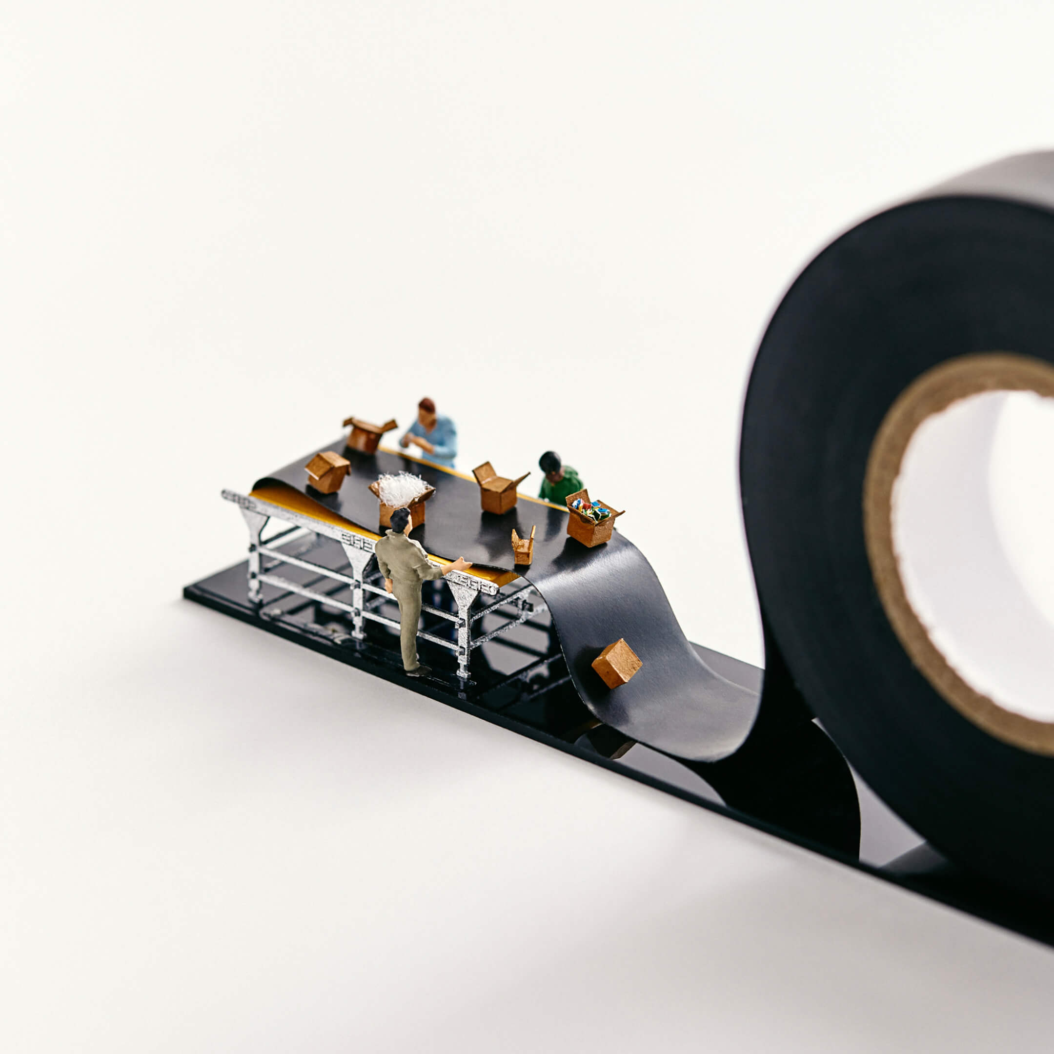 Miniature people pack boxes on a conveyor belt that is actually black electrical tape