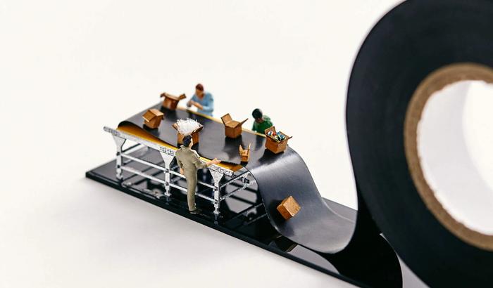 Miniature people pack boxes on a conveyor belt that is actually black electrical tape