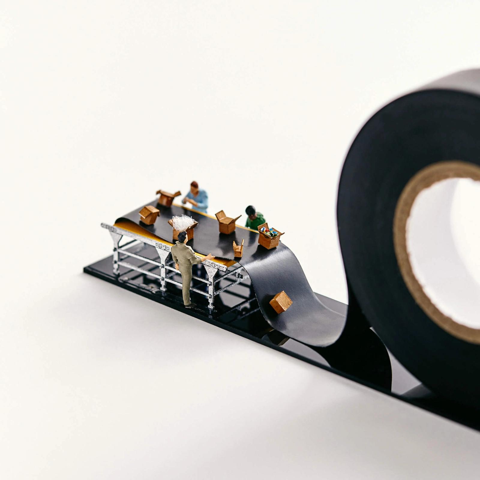 Miniature people pack boxes on a conveyor belt that is actually black electrical tape