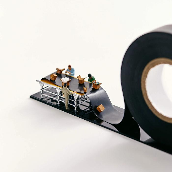 Miniature people pack boxes on a conveyor belt that is actually black electrical tape