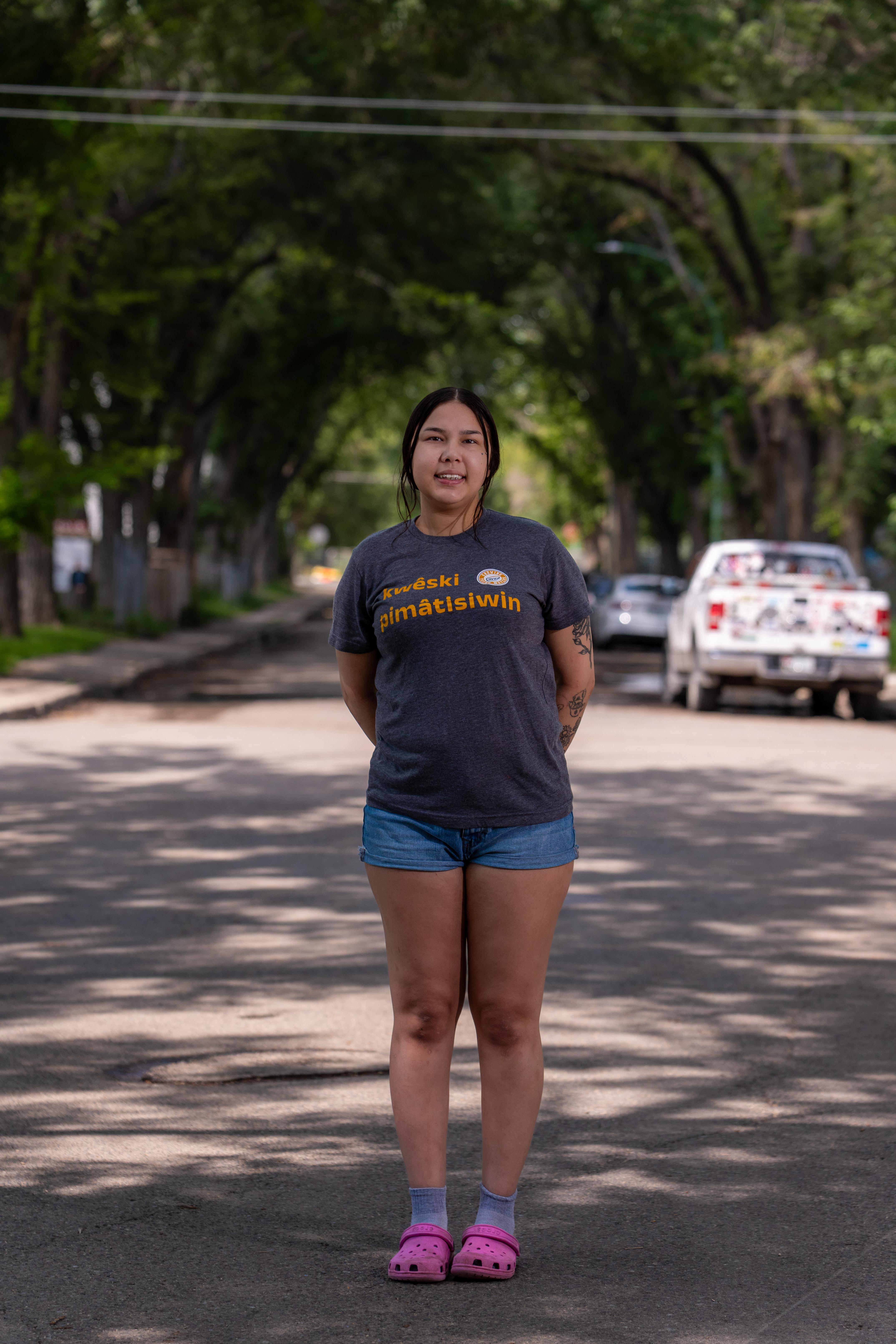 Tyneesha Eaglechild stands in a street, shaded by trees.