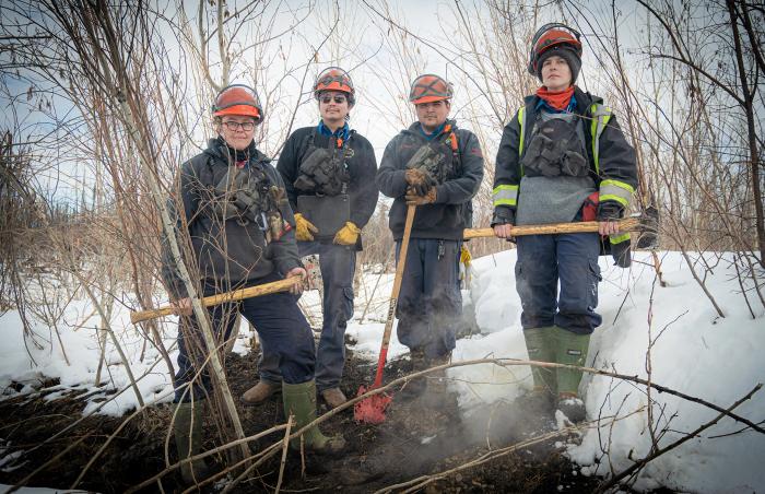 A group shot of the team hunting zombie fires in Canada, showing four people in wildfire fighting gear, From left to right: Sonja Leverkus, Ecosystem Scientist, Senior Fire Lead, Northern Fire WoRx (NFWRx); Donovan Bertrand, Crew Leader NFWRx, member of Acho Dene Koe; David Wilmsmeier, Crew Leader NFWRx, member of Prophet River First Nation; Alexis Jorgensen, Ecologist, NFWRx, Fort Nelson, B.C.