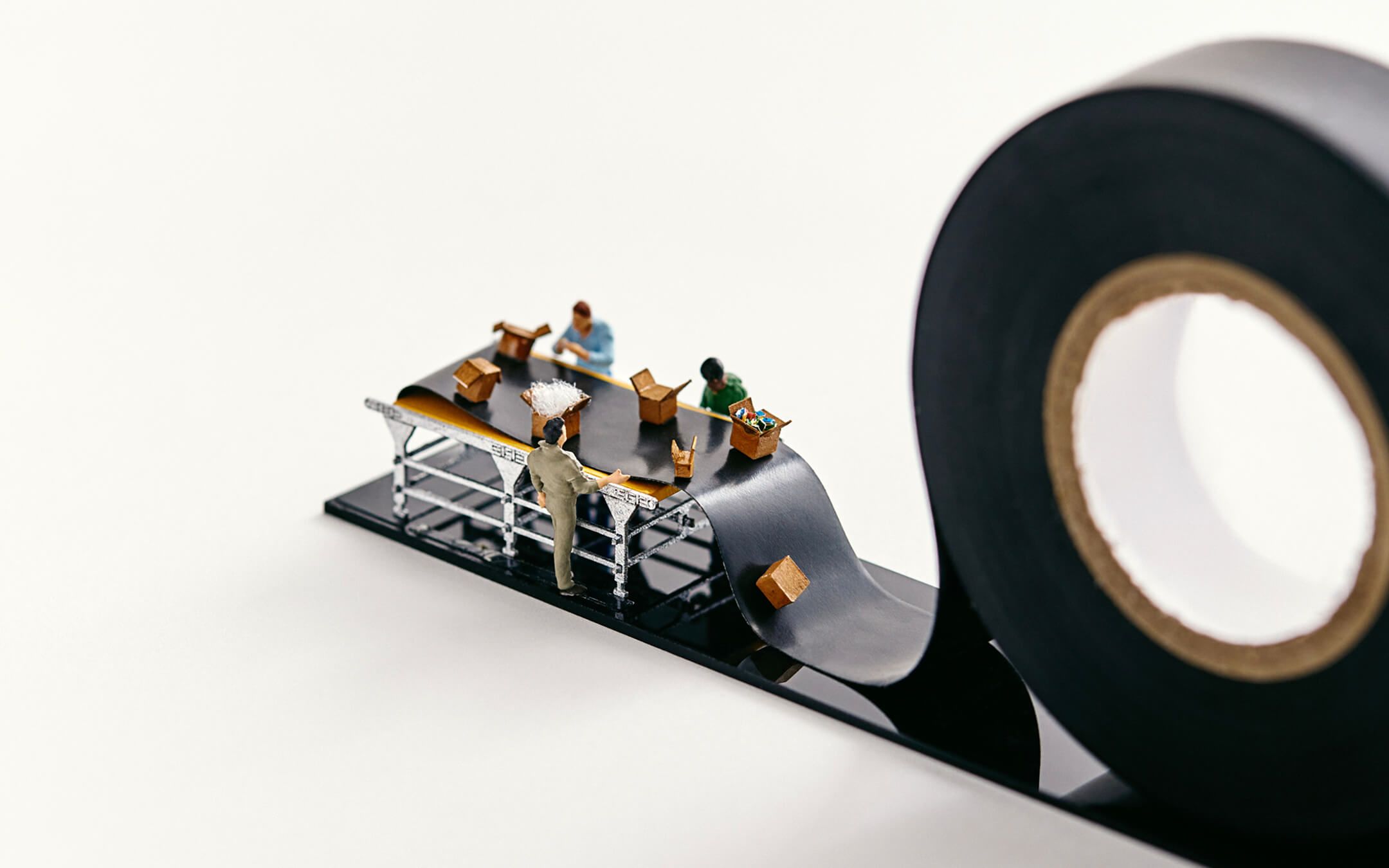 Miniature people pack boxes on a conveyor belt that is actually black electrical tape