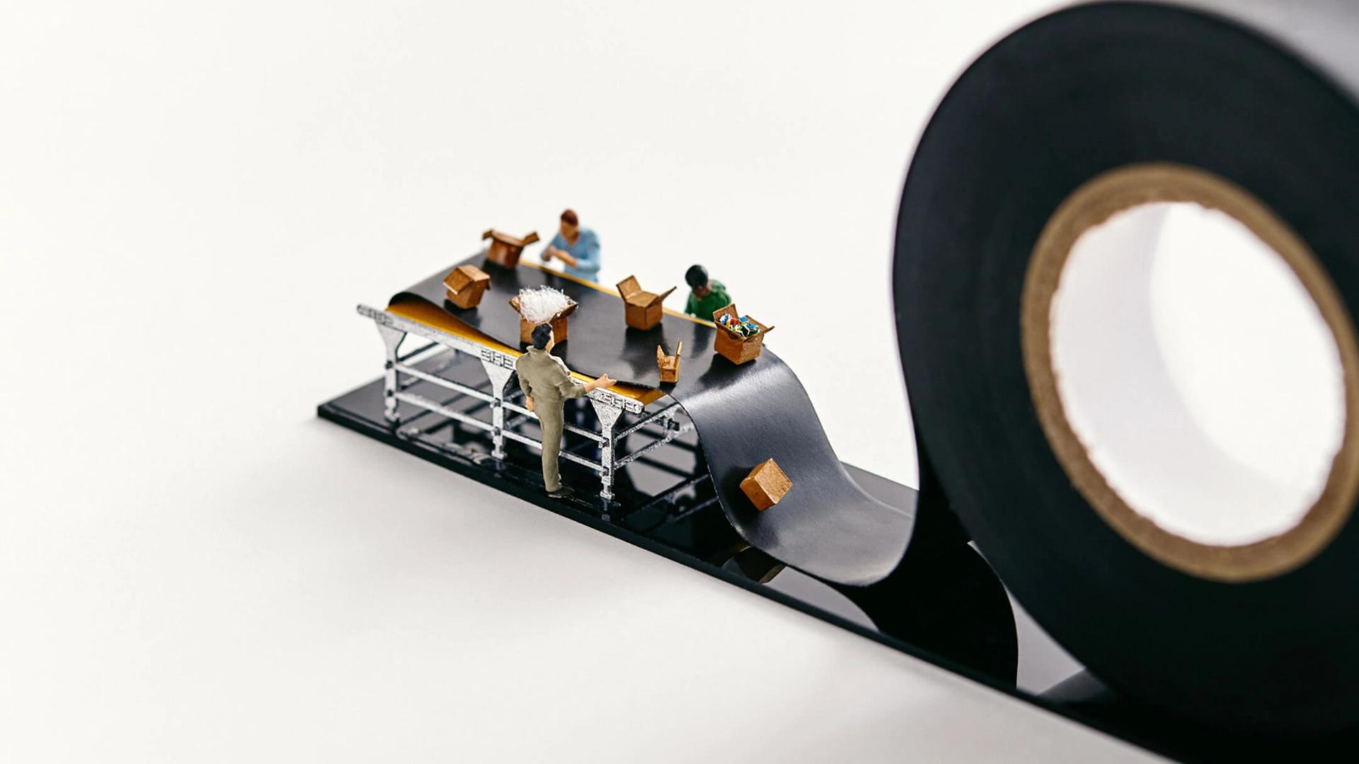 Miniature people pack boxes on a conveyor belt that is actually black electrical tape