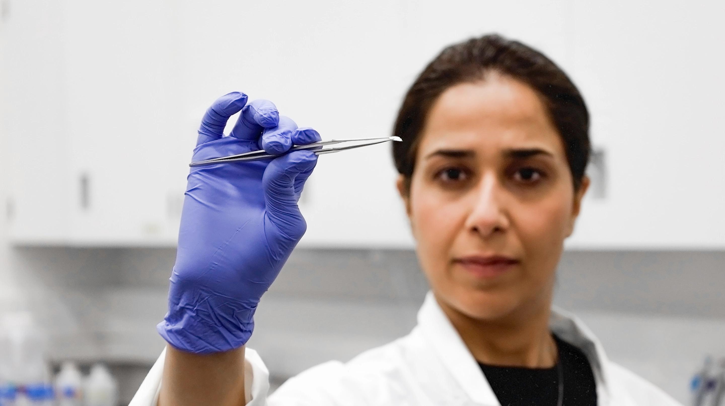 A portrait of Mahla Poudineh wearing gloves and holding tweezers that hold her co-creation, the the Wearable Aptalyzer, a painless wearable device that can track molecules in the body in real time