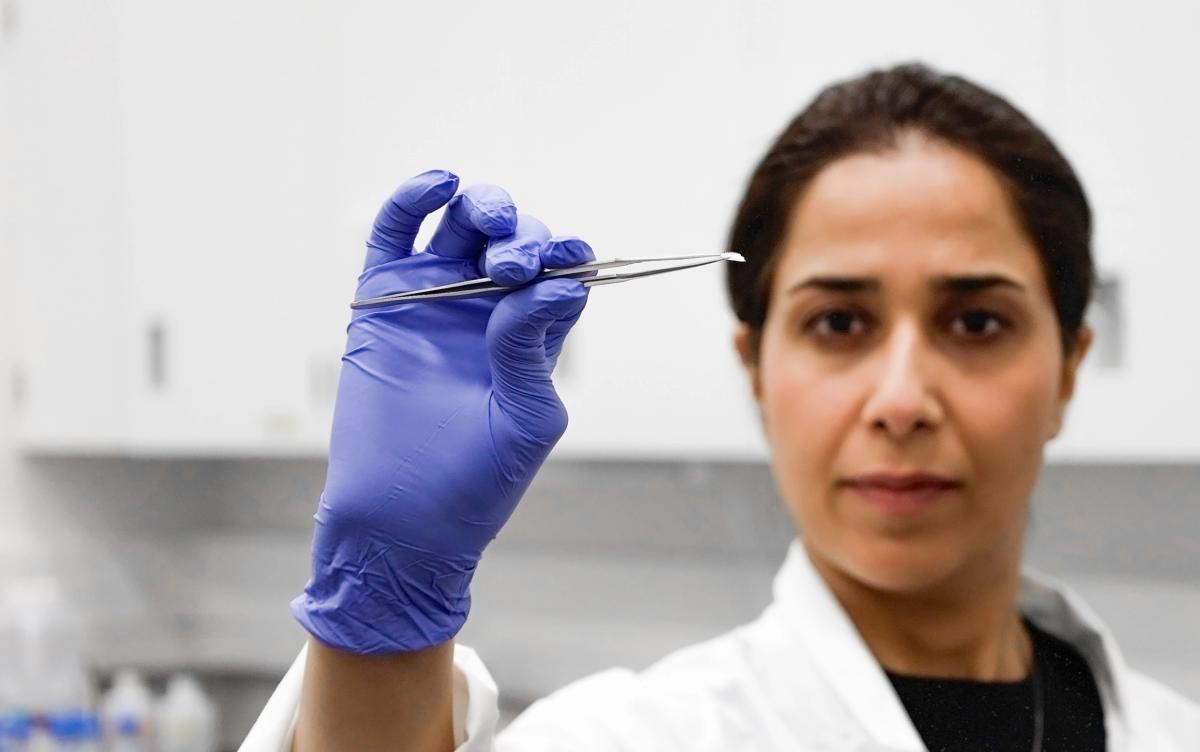 A portrait of Mahla Poudineh wearing gloves and holding tweezers that hold her co-creation, the the Wearable Aptalyzer, a painless wearable device that can track molecules in the body in real time