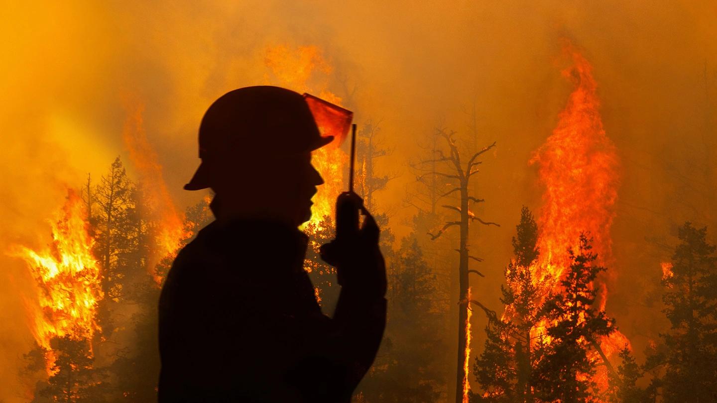 A silhouette of a firefighter standing in front of a burning, smoke-filled forest. The firefighter is talking into their walkie-talkie