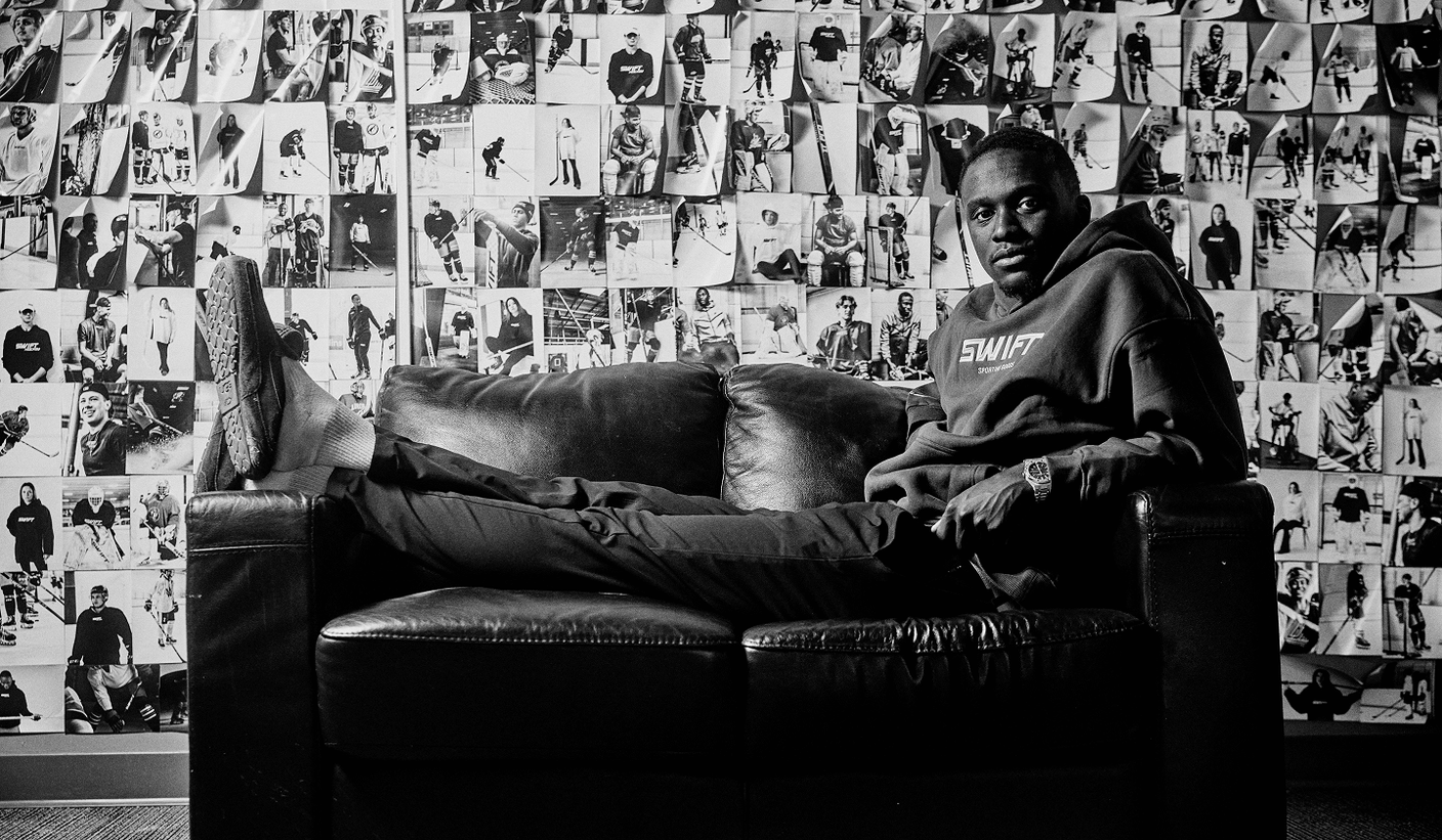 Founder of Swift Hockey Zech Thomas sitting on a couch with a wall covered with hockey images of his budget hockey stick company.