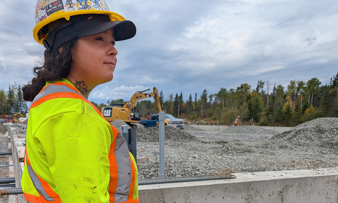 A person in construction gear and a hardhat over a baseball cap wearing a bright yellow and orange reflective high-visibility vezt looks out over a construction site