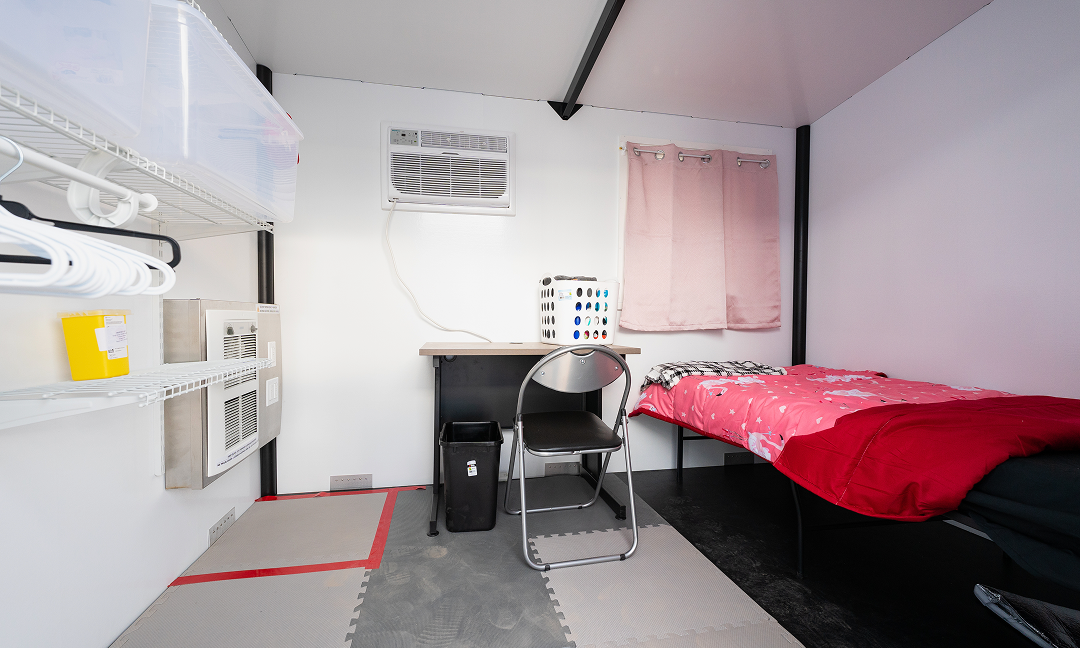 The interior of a small prefabricated shelter at the the Village at Pine Tree Park on the outskirts of Sydney on Cape Breton Island, showing a bed with a red bedspread, a small desk with a chair and shelves for storage.