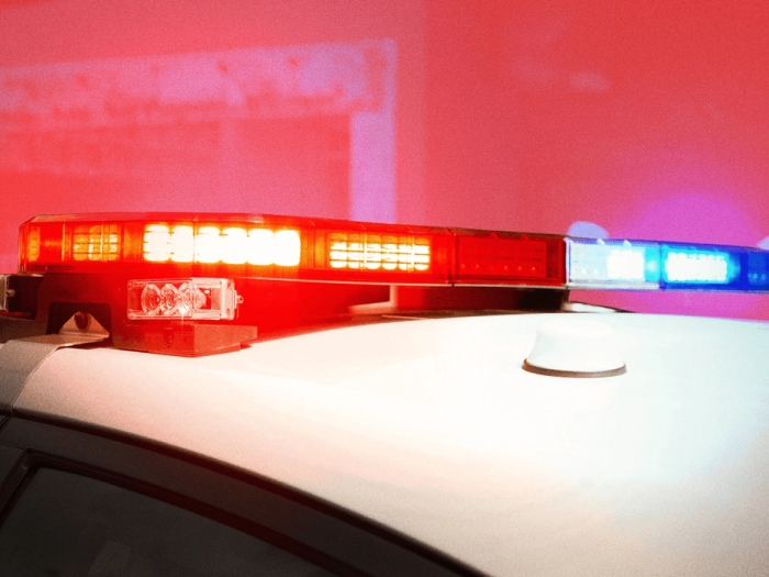 A photo illustration of a flashing police vehicle siren, shown in closeup on top of a white vehicle, against a reddish illustration background, for a piece on AI use for 911 calls in Canada