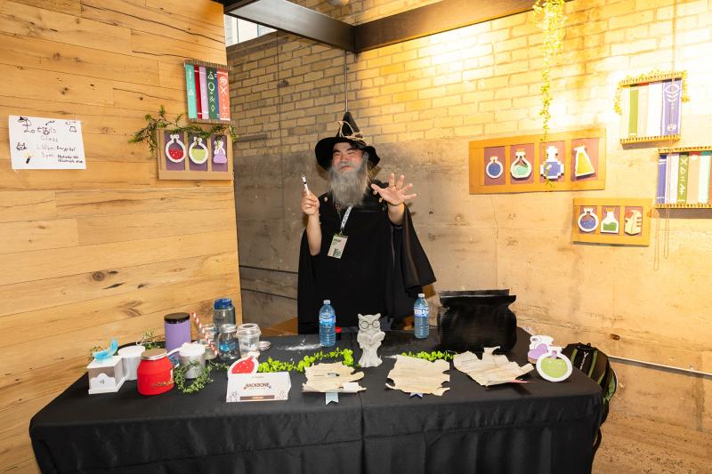 Daniel Guo wears a wizard costume with a peaked hat and a wand and makes a gesture as if performing a spell behind a wizard-themed booth to show off his homemade energy supplement BackBone at the 2026 Socratica Symposium in Kitchener, Ontario Canada