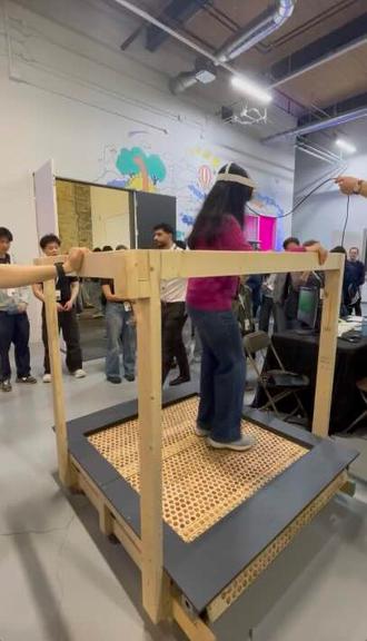 A person wearing a VR headset walks on an omnidirectional treadmill framed with a wood frame while someone holds the cord of her vr headset and onlookers look on at the Socratica symposium