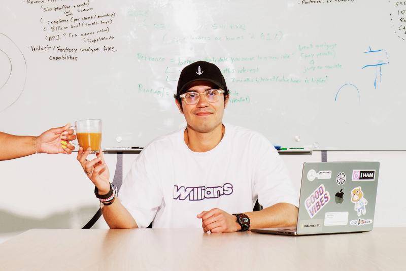 Swish Goswami sitting in a board room with a whiteboard in the background, being handed a coffee, with a laptop beside him on a desk
