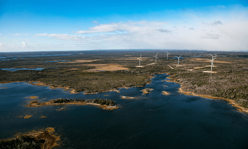 An aerial view of the site of Spaceport Nova Scotia near Canso, Nova Scotia showing dark blue ocean and green shorelines with wind turbines in the distance then blue sky and clouds
