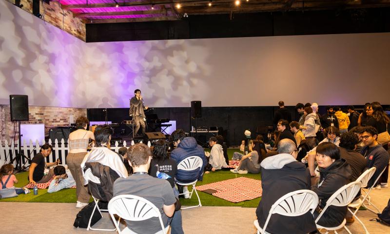 Wesley Ou, holding a microphone, performs a hyperpop set on the Socratica Symposium stage while onlookers in white chairs or sitting on fake grass and checked picnic blankets look on