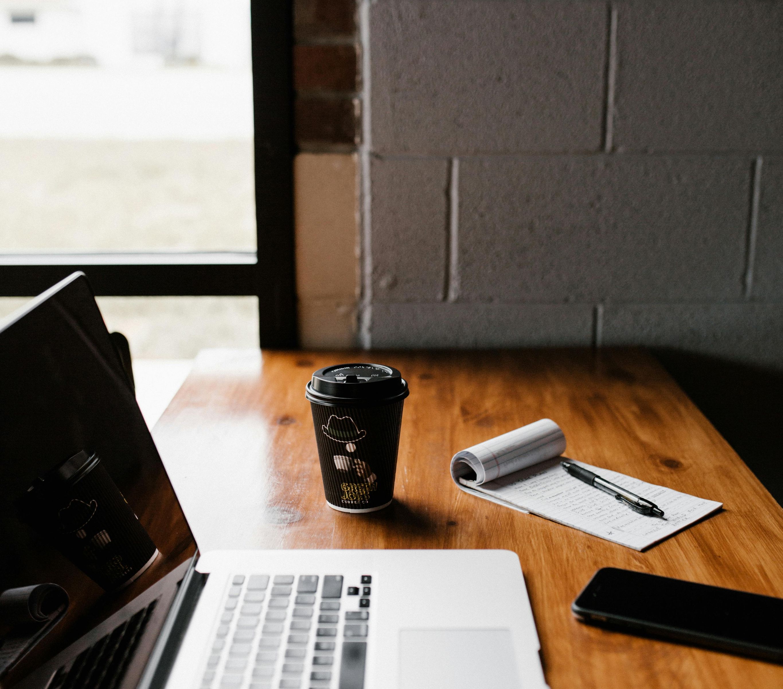 Laptop next to a coffee cup, notepad and a mobile phone