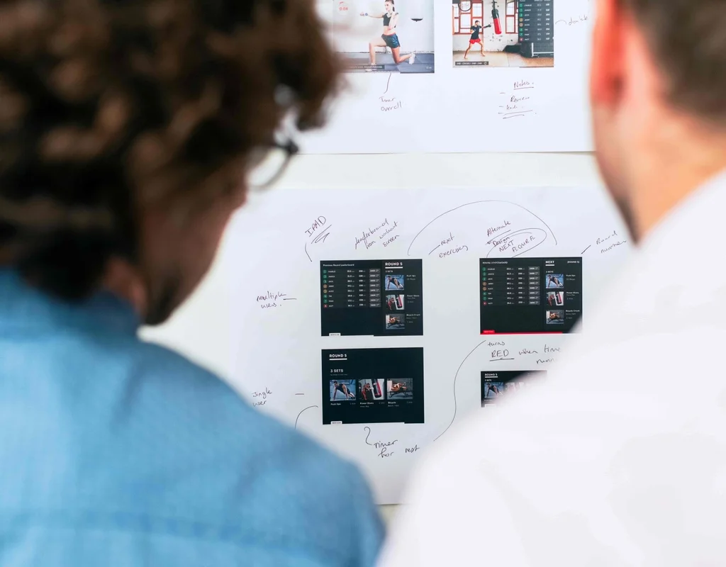 Two people discussing application designs printed out and hanged on a wall