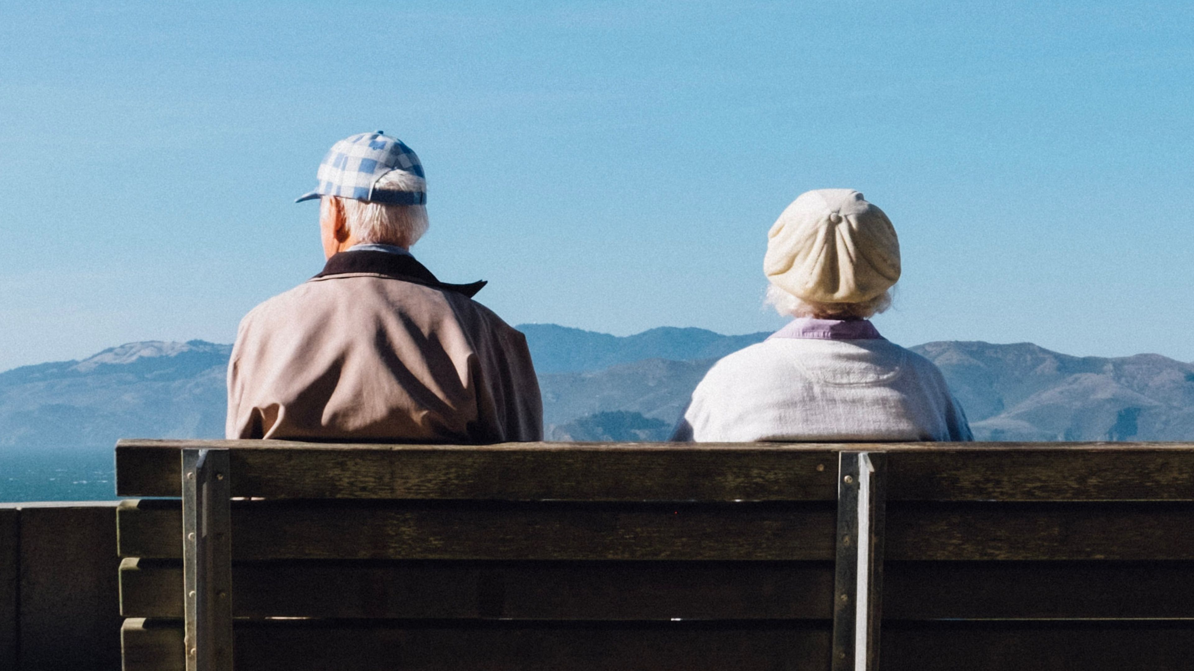The backs of an older woman and man as they sit on a bench taking in the views of mountains.  