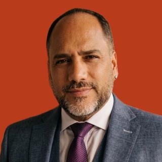 Isra Barriga, in a grey suit and purple tie against a rust background.