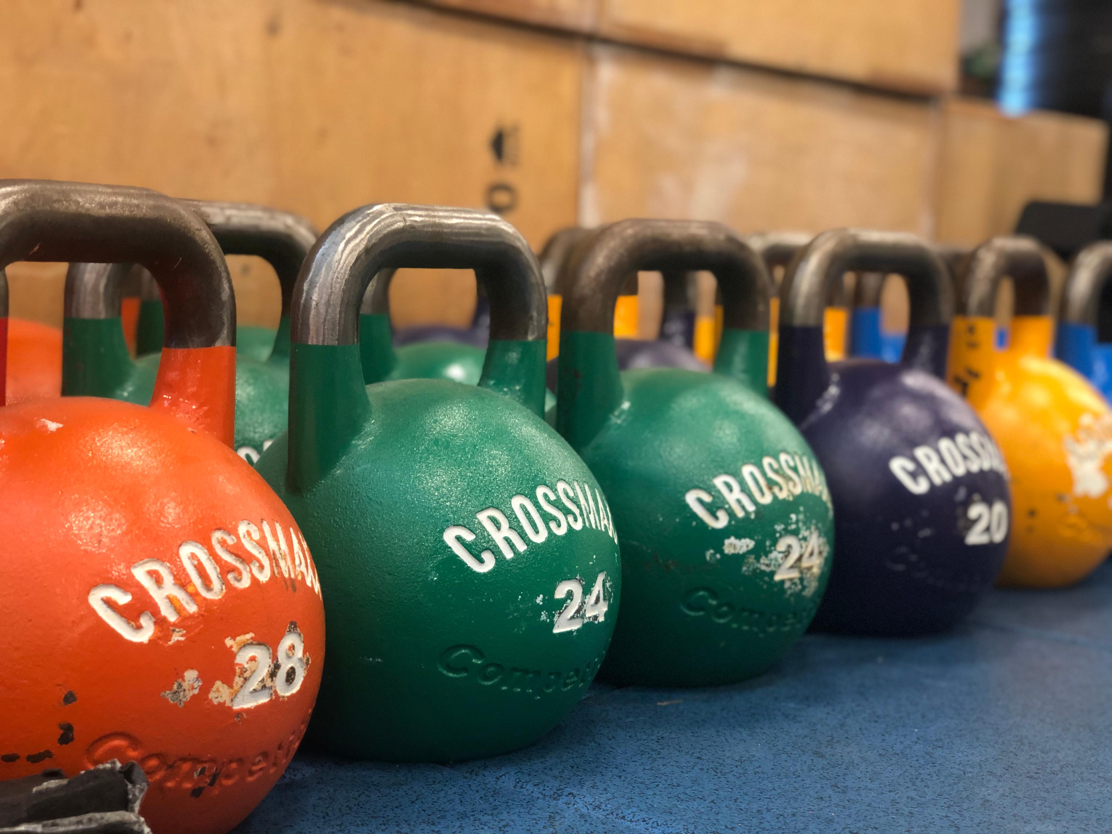 Verschillende kettlebells in verschillende kleuren (oranje, groen, geel, blauw) netjes op een rij geplaatst op een blauwe vloer.