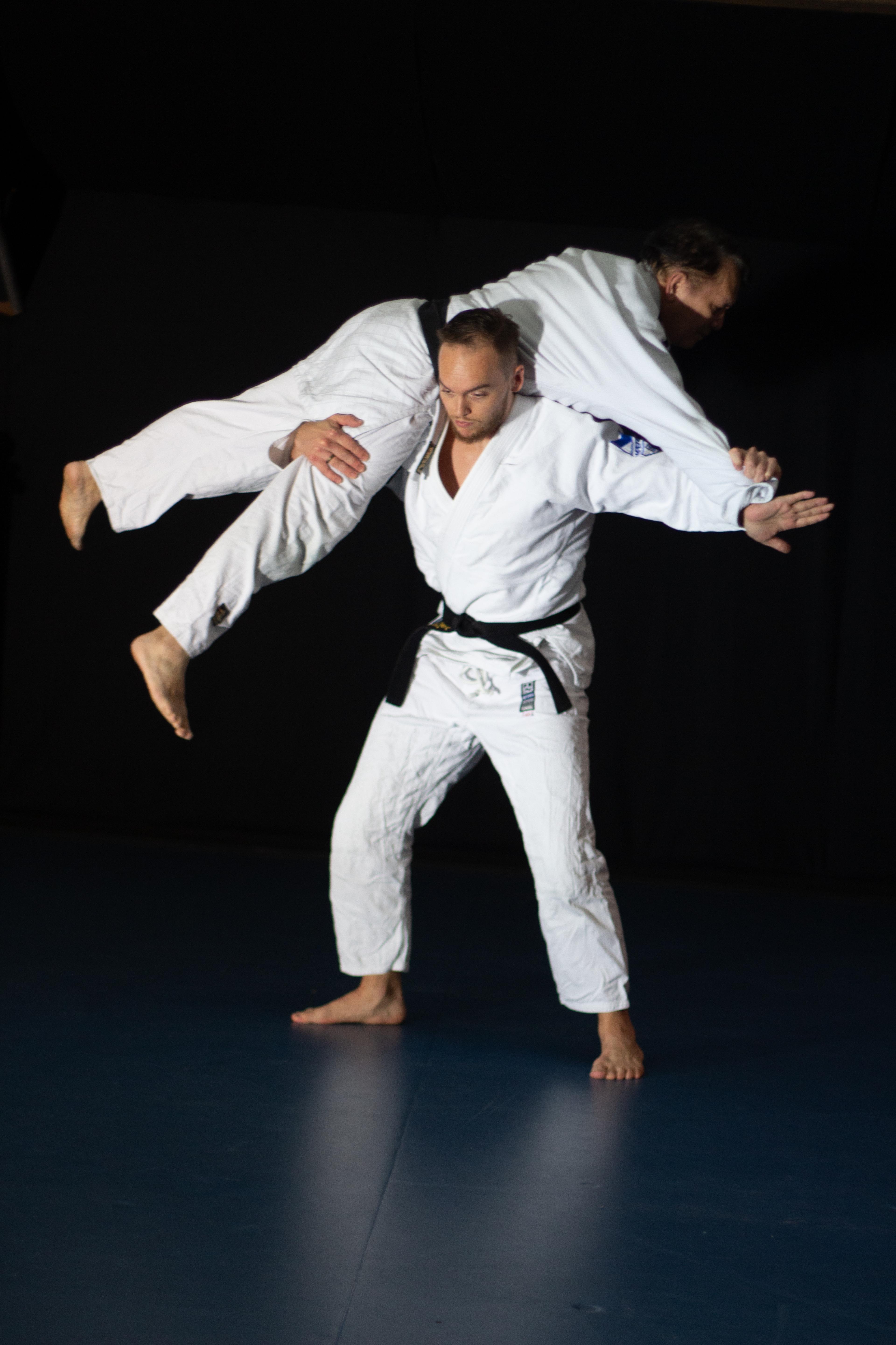 Twee mensen in judo-pakken oefenen een worp in een donkere kamer.