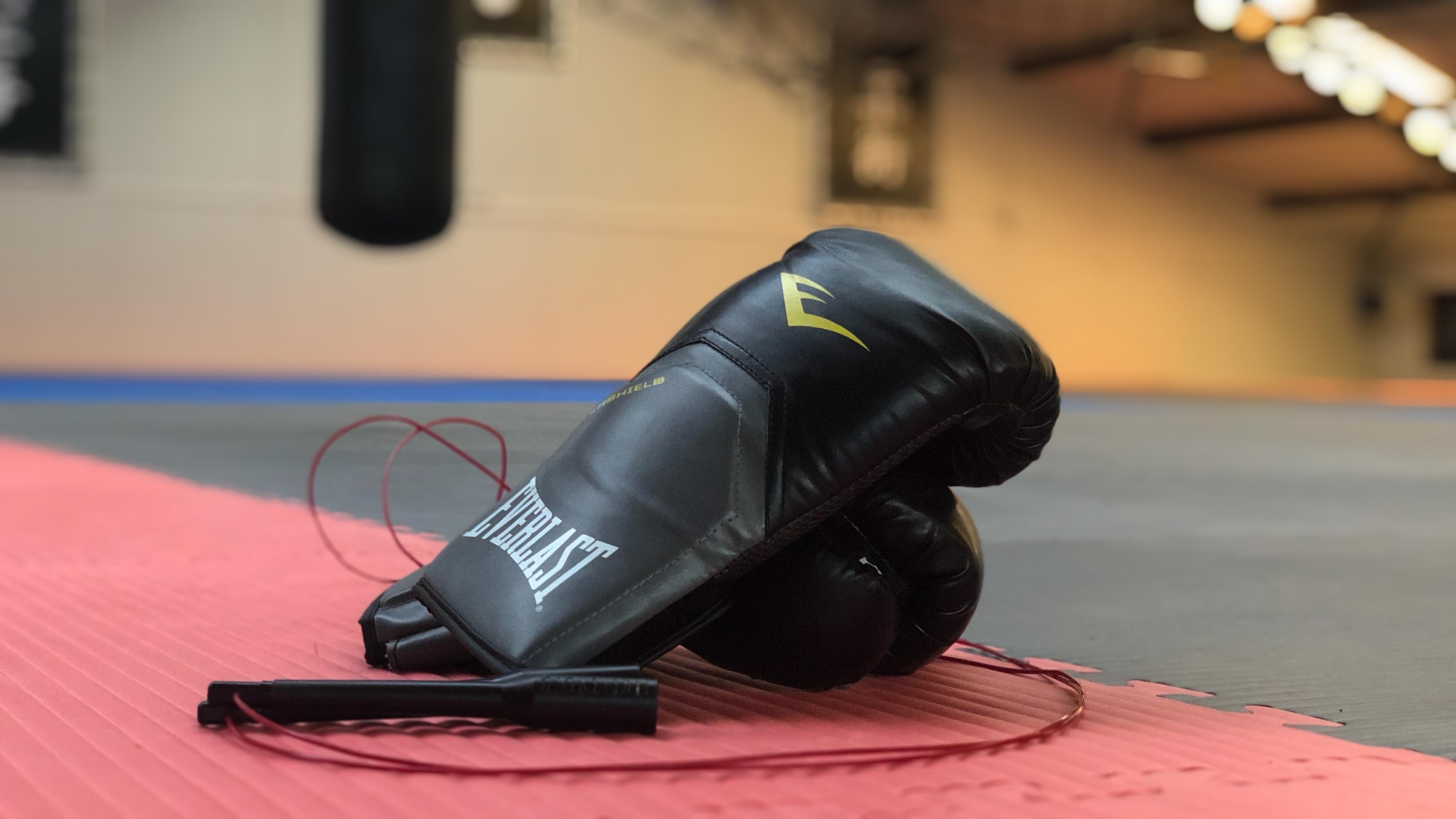 Bokshandschoenen en een springtouw liggen op een rode mat in een dojo of trainingsruimte.